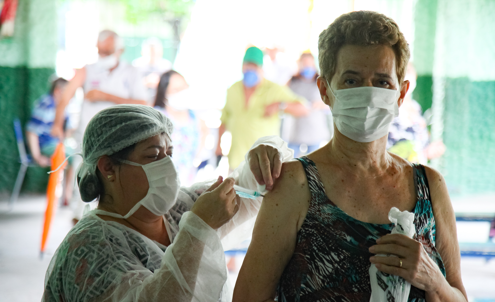 mulher profissional de saúde aplicando vacina em idosa que usa máscara e olha para a foto